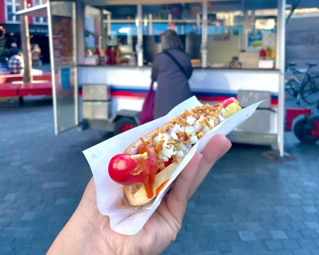 Agalya holding a Pølsemand, a Danish hotdog in front of street cart